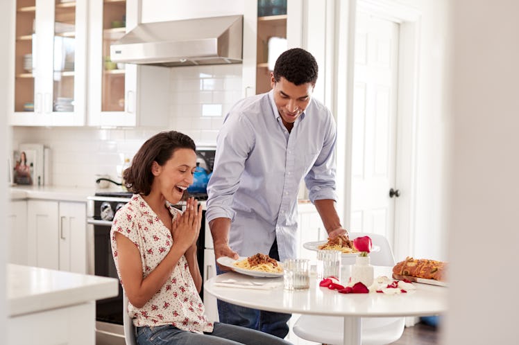 Young African American adult woman sitting at the table in the kitchen, her partner surprising her b...