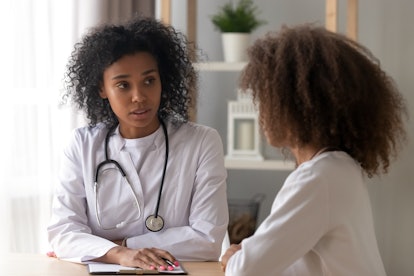 Serious African American female doctor talk to teenage girl sitting at table, focused black woman ph...