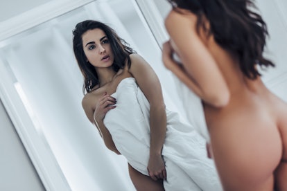 Sexy naked woman covered by blanket is looking in mirror in bedroom