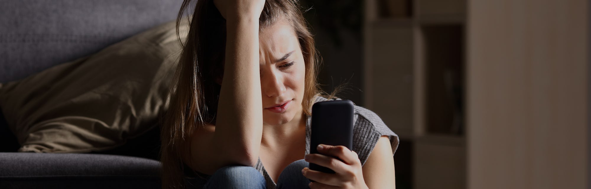 Front view of a sad teen checking phone sitting on the floor in the living room at home with a dark ...