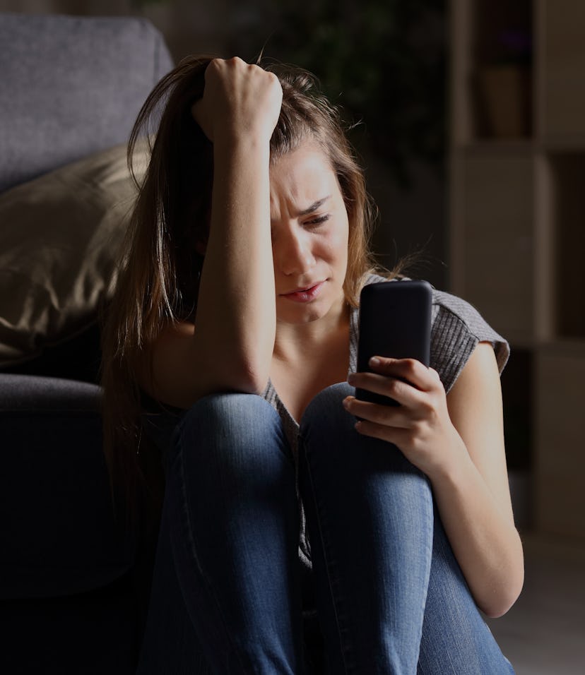 Front view of a sad teen checking phone sitting on the floor in the living room at home with a dark ...