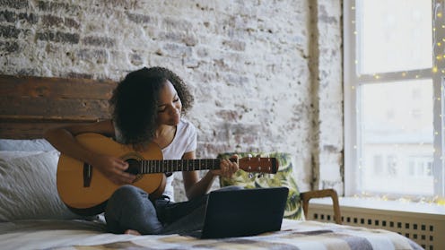 Curly african american teenager girl learning to play guitar using laptop computer sitting on bed at...