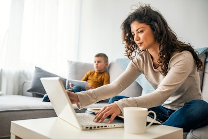 Smiling mom working at home with her child on the sofa while writing an email. Young woman working f...