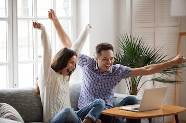 A happy couple celebrates together in front of their laptop with their arms' raised. 