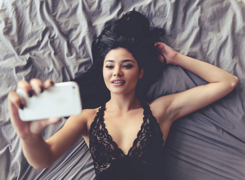 Top view of sensual young woman in black lingerie making selfie using a smartphone while lying on be...