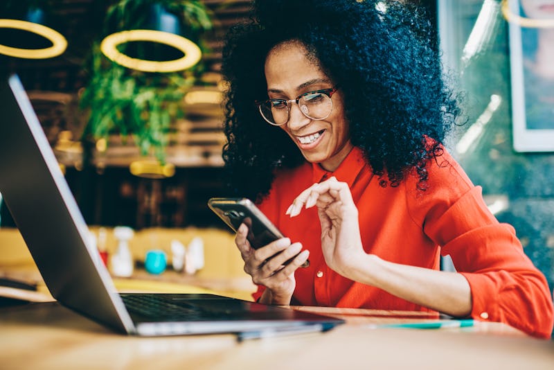 Cheerful African American young woman laughing while reading funny notification on smartphone sittin...