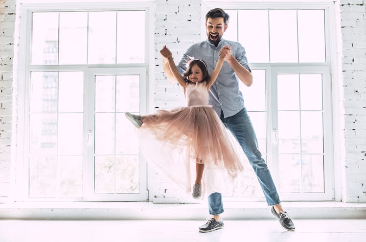 A younger father dressed in jeans and a button-down shirt laughs while he lifts his ballerina daught...