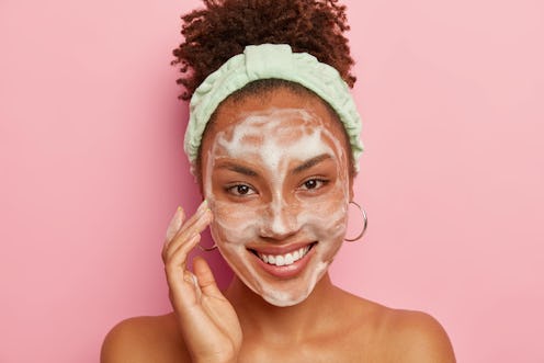 Happy dark skinned woman pampers face with bubble cleansing foam, has hygienic treatments in bathroo...