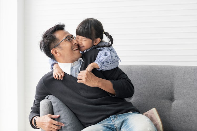 An Asian father and daughter laugh while snuggling on the couch in their living room.