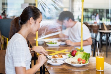Asian man and woman sitting separated in restaurant eating food with table shield plastic partition ...