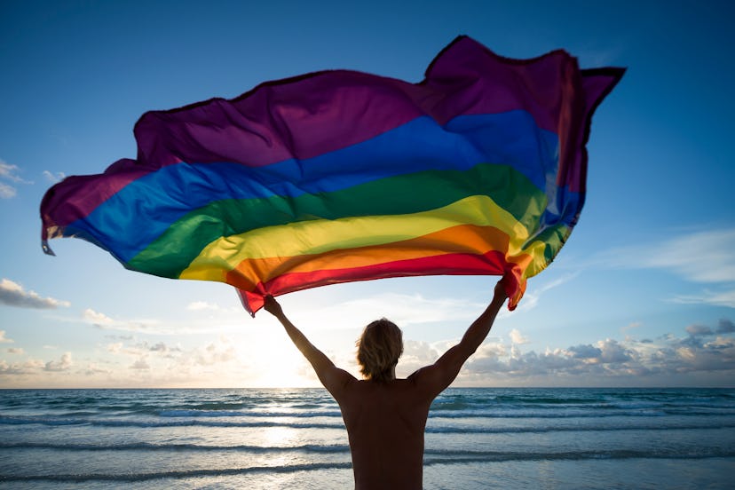 Silhouette of man holding a gay pride rainbow flag fluttering against the rising sun on an empty bea...