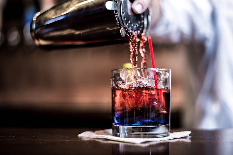 A red cocktail being poured from a shaker into a glass on a napkin on a bar top