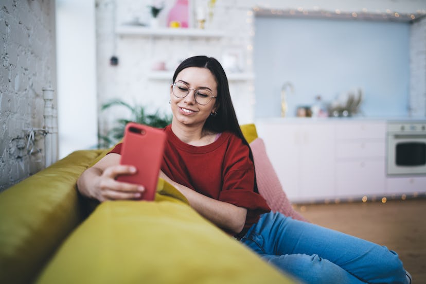 Young woman in casual clothing and glasses talking on video call on red mobile phone while sitting o...