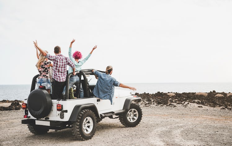 A group of friends in a Jeep Wrangler throw their hands in the air while parked near the shore.