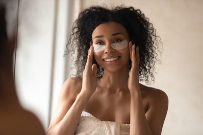 A woman in a bath towel puts under-eye patches while working on her skincare.
