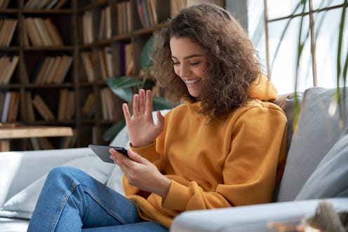 Happy young hispanic latin teen girl sit on sofa at home holding phone looking at screen waving hand...