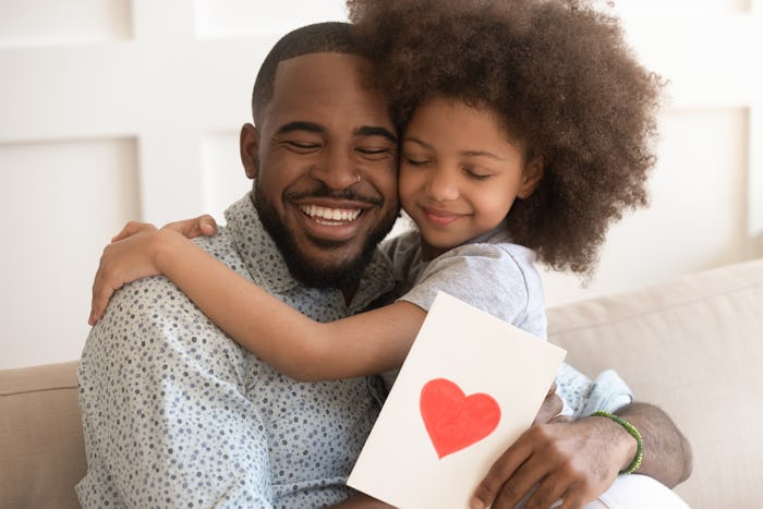 Happy affectionate african american dad embracing little child daughter holding greeting card with r...