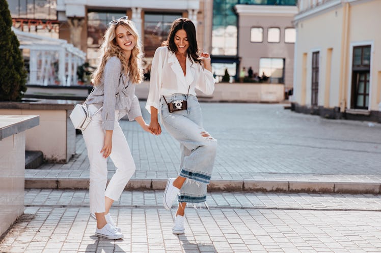 Two friends wearing jeans, trendy tops, and white sneakers hold hands and strike a pose on a sidewal...