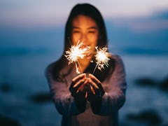 Young happy woman stands on beach with sparkler in sunset light.