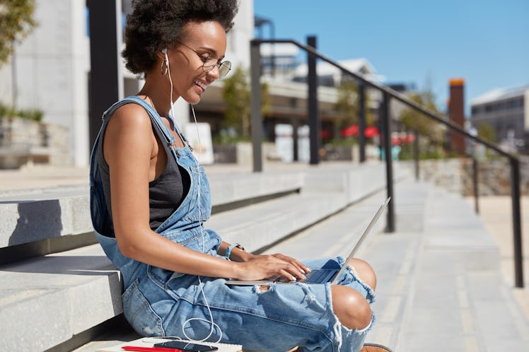 Sideways shot of black satisfied young woman checks email box on laptop computer, reads news, wears ...
