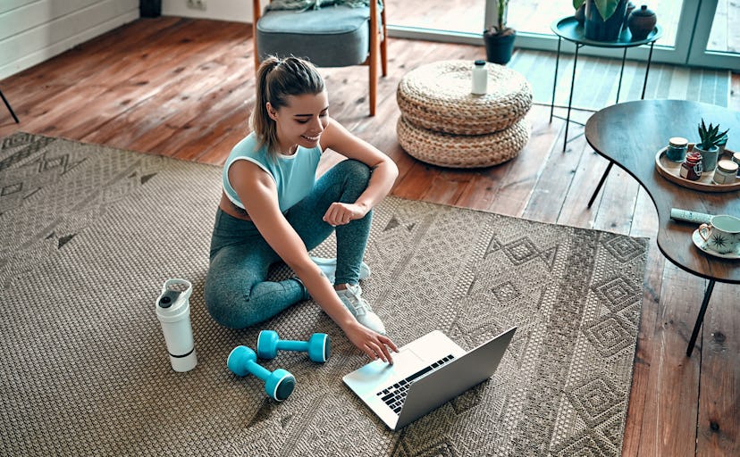 A sporty woman in sportswear is sitting on the floor with dumbbells and a protein shake or a bottle ...