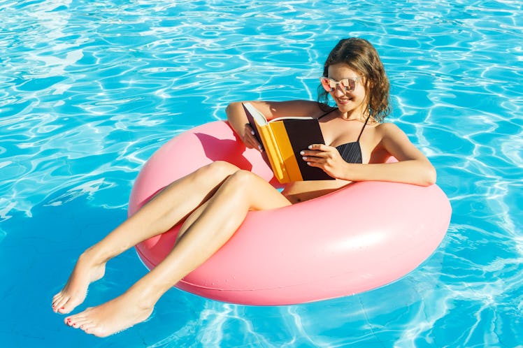 A happy girl in a bikini is floating in the pool on a pink tube with a book in her hand.