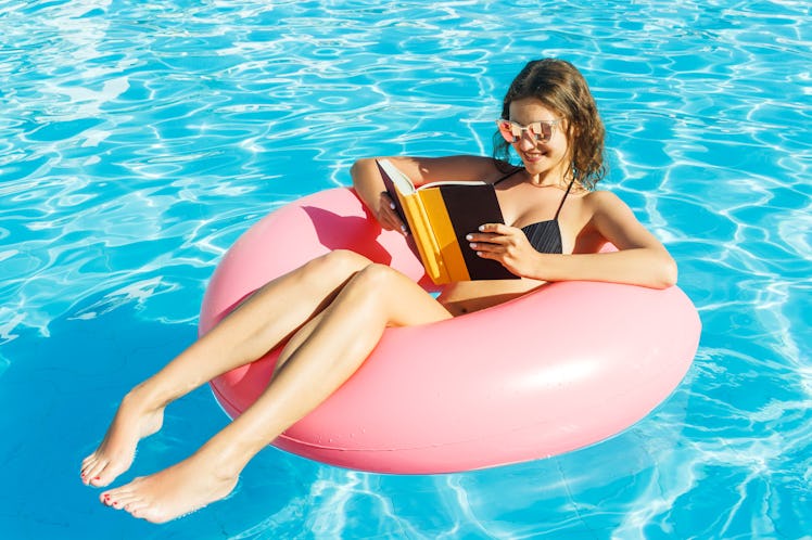 A happy girl in a bikini is floating in the pool on a pink tube with a book in her hand. 