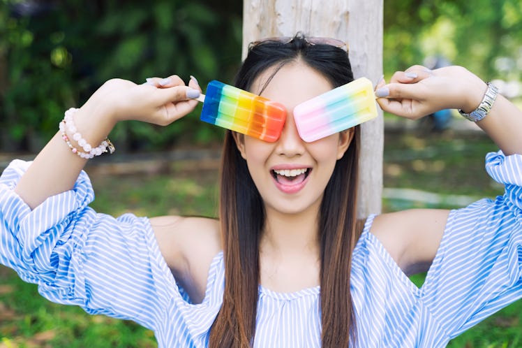 A woman smiles while holding up popsicles to her eyes in the summer. 