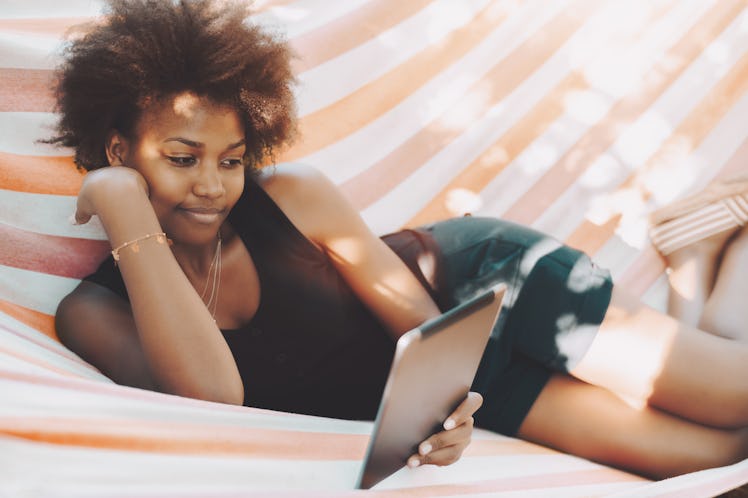 Beautiful relaxed black female student with curly African hair is laying on striped hammock in campu...