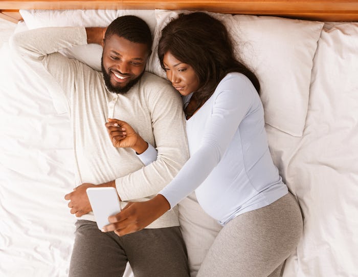 Married pregnant black couple taking selfie on smartphone while resting on bed at home, top view