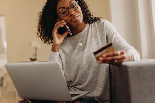 Woman sitting on sofa at home talking on mobile phone holding a credit card with a laptop on her lap...