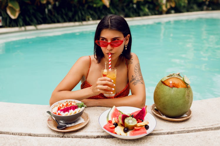 Confident brunette tan woman in red cat eyes sunglasses relaxing in pool with plate of exotic ...