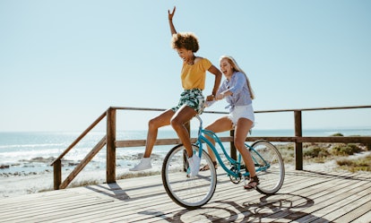 Two friends laugh while riding a bike on a boardwalk along the beach in the summer.
