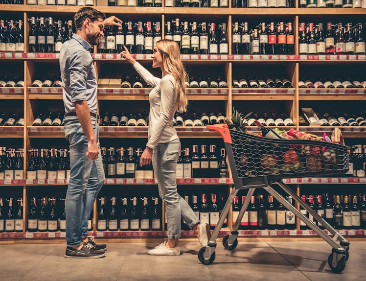 A trendy couple smiles while choosing wine in a liquor store.
