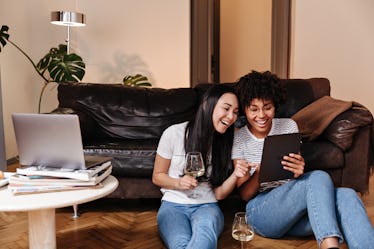 Two best friends sit on the floor with glasses of wine, while looking at videos on their tablet comp...