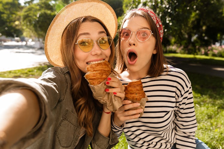 Two happy young girls friends having fun at the park, taking a selfie, holding croissants