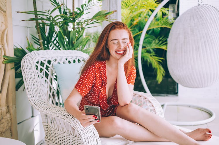 A happy woman in a red polka dot romper sits on her front porch on a white wicker chair holding her ...