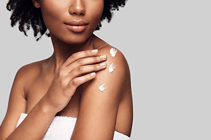 Beauty routine. Close up of young African woman applying body cream and smiling while standing again...