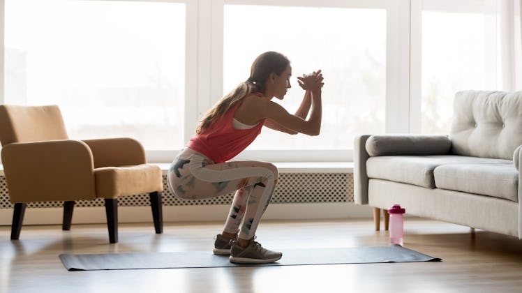 Sporty young woman doing squat morning exercise alone in living room, serious fit girl wearing sport...
