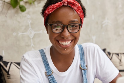 Headshot of pleasant looking young woman has shining smile, wears piercing in nose and lips, spectac...