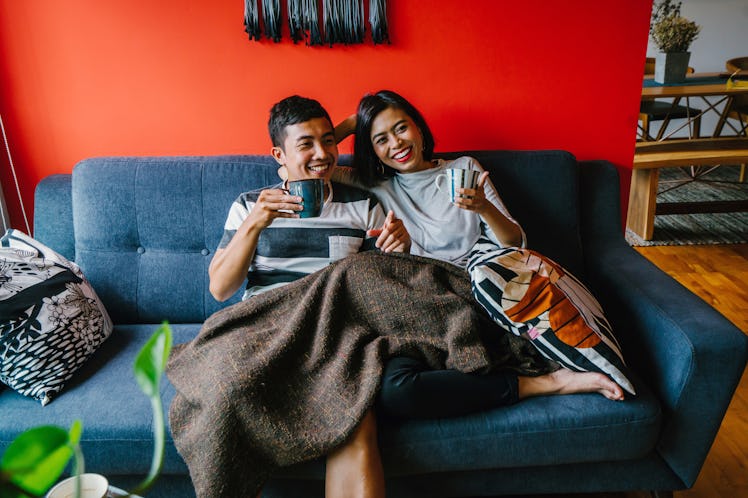 A young Asian couple laughs and cuddles on the couch under a blanket.