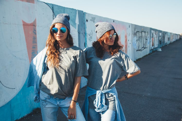 Two girls wearing gray T-shirts, gray beanies, and blue sunglasses stand in front of a wall outside.