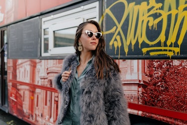 A stylish woman wears sunglasses, a gray button-down, and a gray faux fur coat.