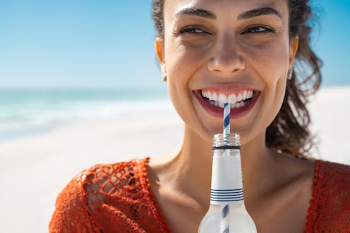 Closeup face of young woman drinking fresh sparkling water from a glass bottle at beach. Portrait of...