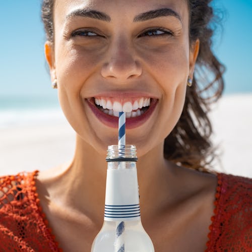 Closeup face of young woman drinking fresh sparkling water from a glass bottle at beach. Portrait of...