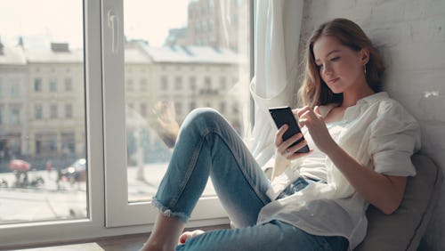 Close up pretty young woman sitting on a windowsill at home and texting on her phone communication f...