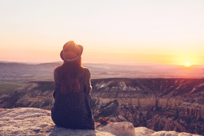 A view from the back of a girl in a hat sits on a mountain and admires the beautiful view and dawn o...