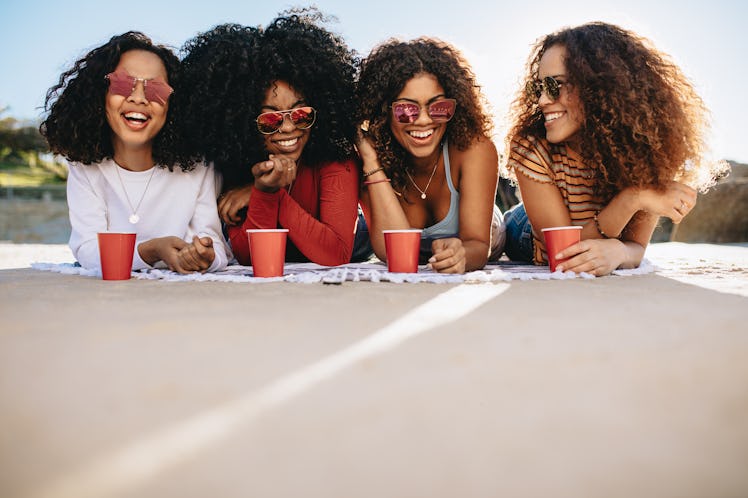A group of friends wearing sunglasses and holding red cups lounge on the ground during a picnic in t...