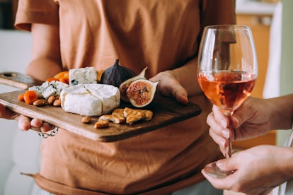 A woman holds a cheese board, filled with snacks, while another set of hands hold a glass of wine. 