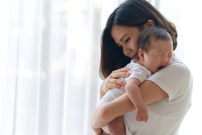 Cute yawning Asian infant baby on mothers hands standing in the room near the window. Mother hugging...